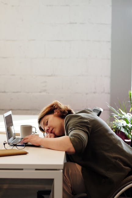 A tired woman in a wheelchair sleeps at her desk in a modern office setting.